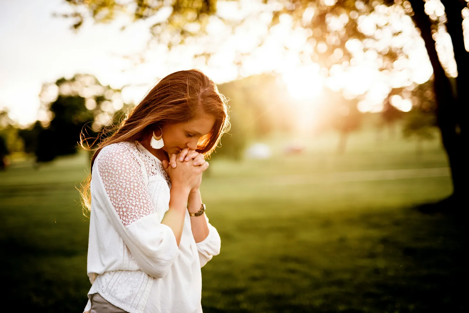A woman standing in a field at sunset with her hands folded in prayer
